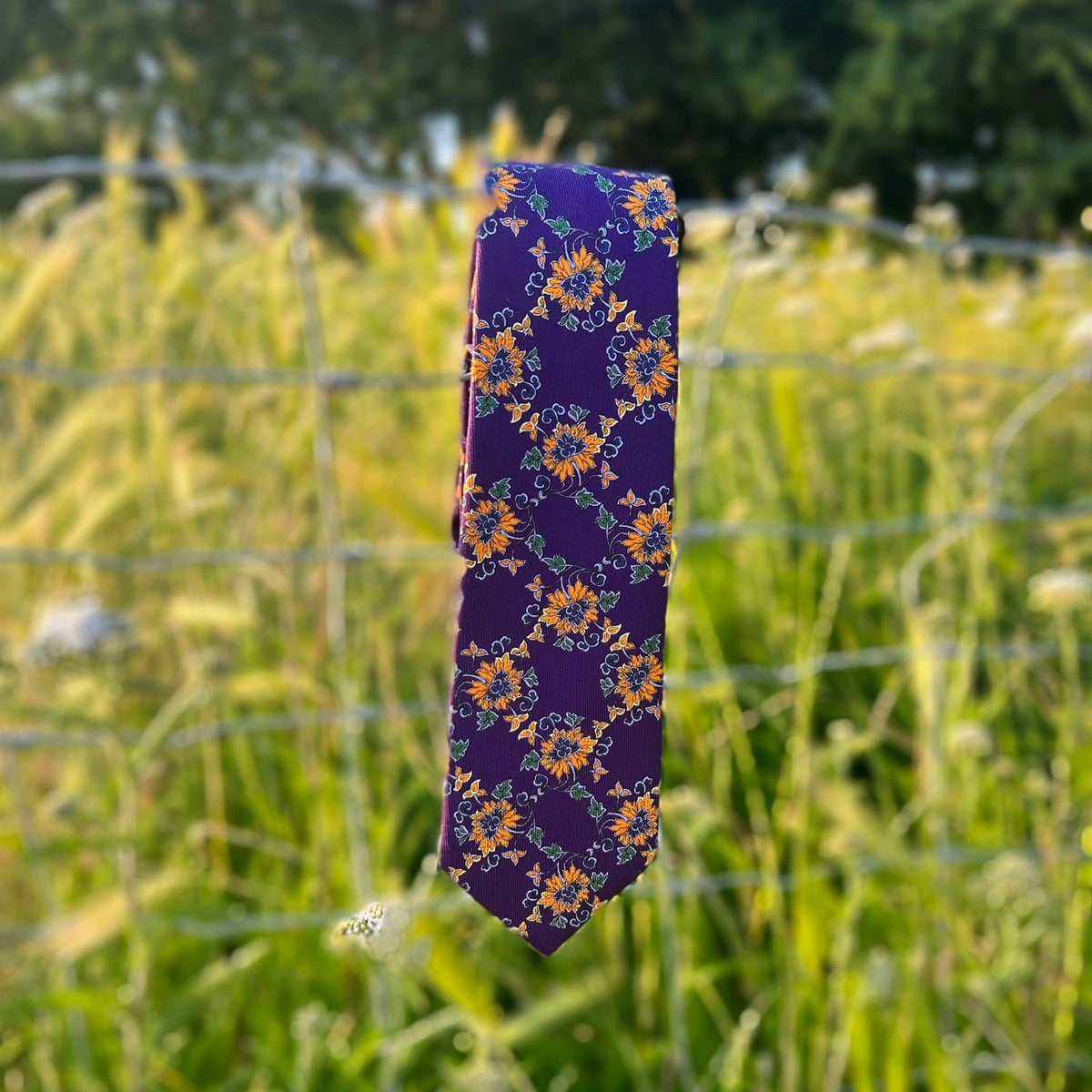 A purple necktie with a yellow and orange floral pattern, hanging outdoors on a wire fence and green grass.