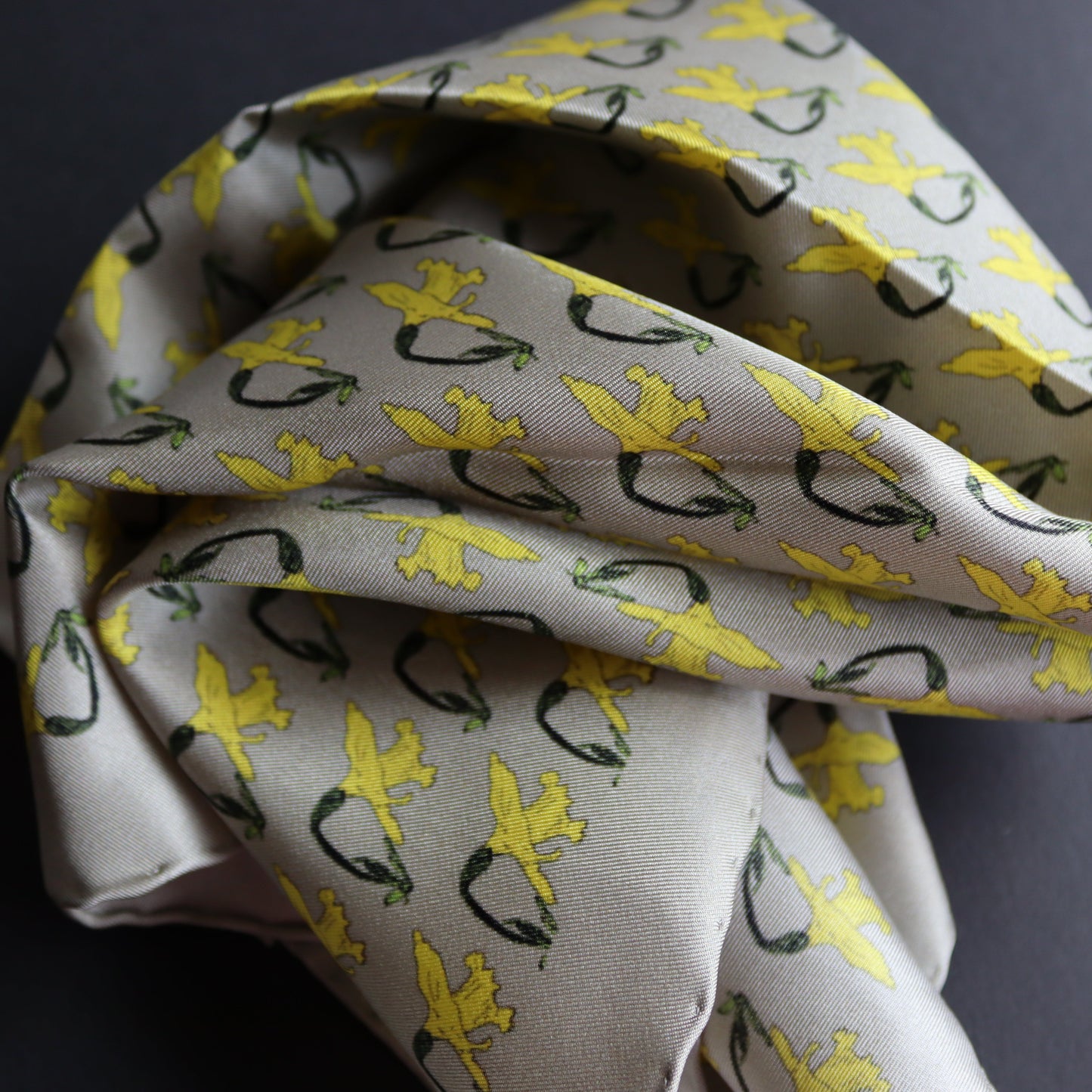 A silk pocket square with a light grey base, featuring a repeating pattern of yellow daffodils, displayed on a dark grey surface.