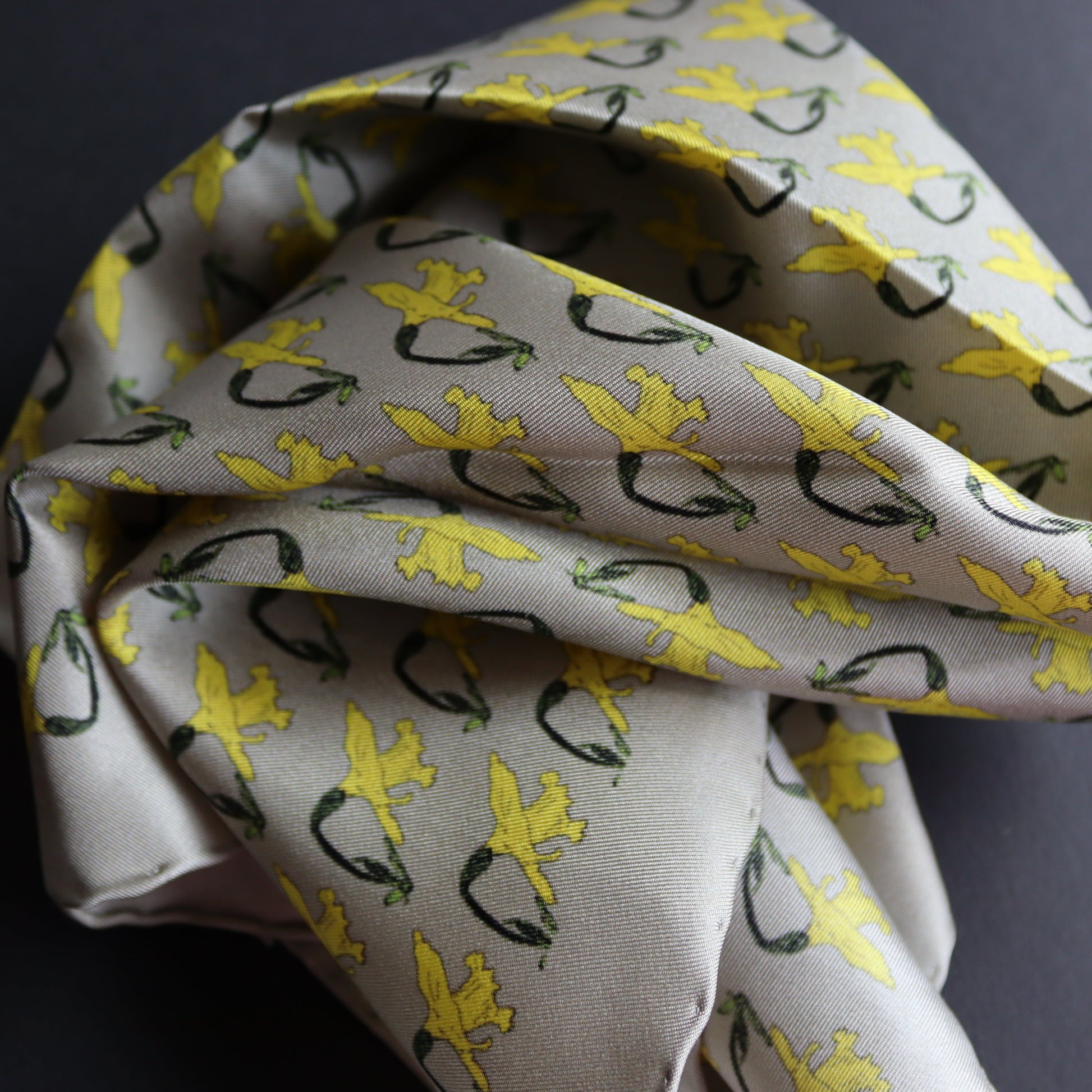 A silk pocket square with a light grey base, featuring a repeating pattern of yellow daffodils, displayed on a dark grey surface.
