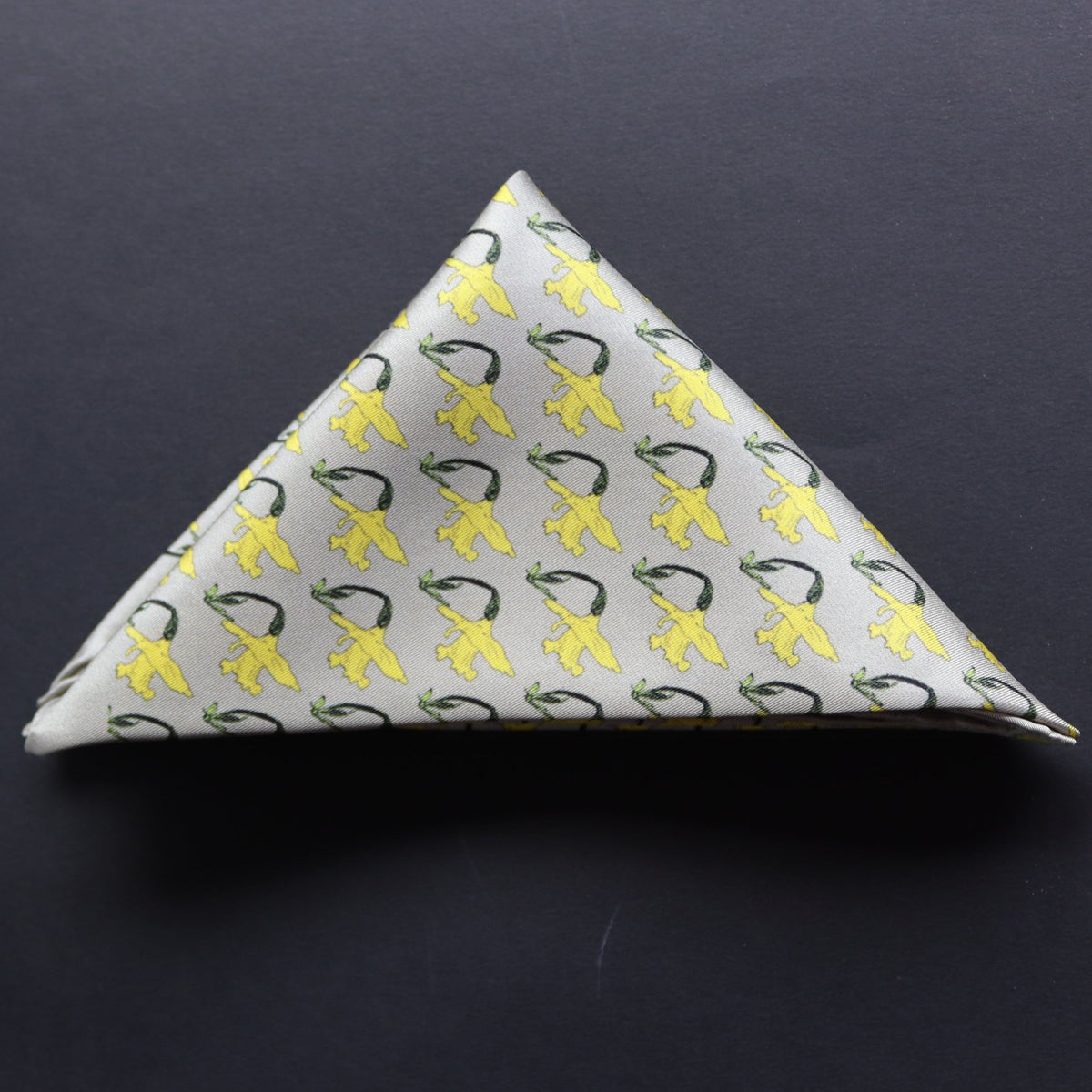 A silk pocket square with a repeating pattern of yellow daffodils on a dusky grey background and folded into a triangle.