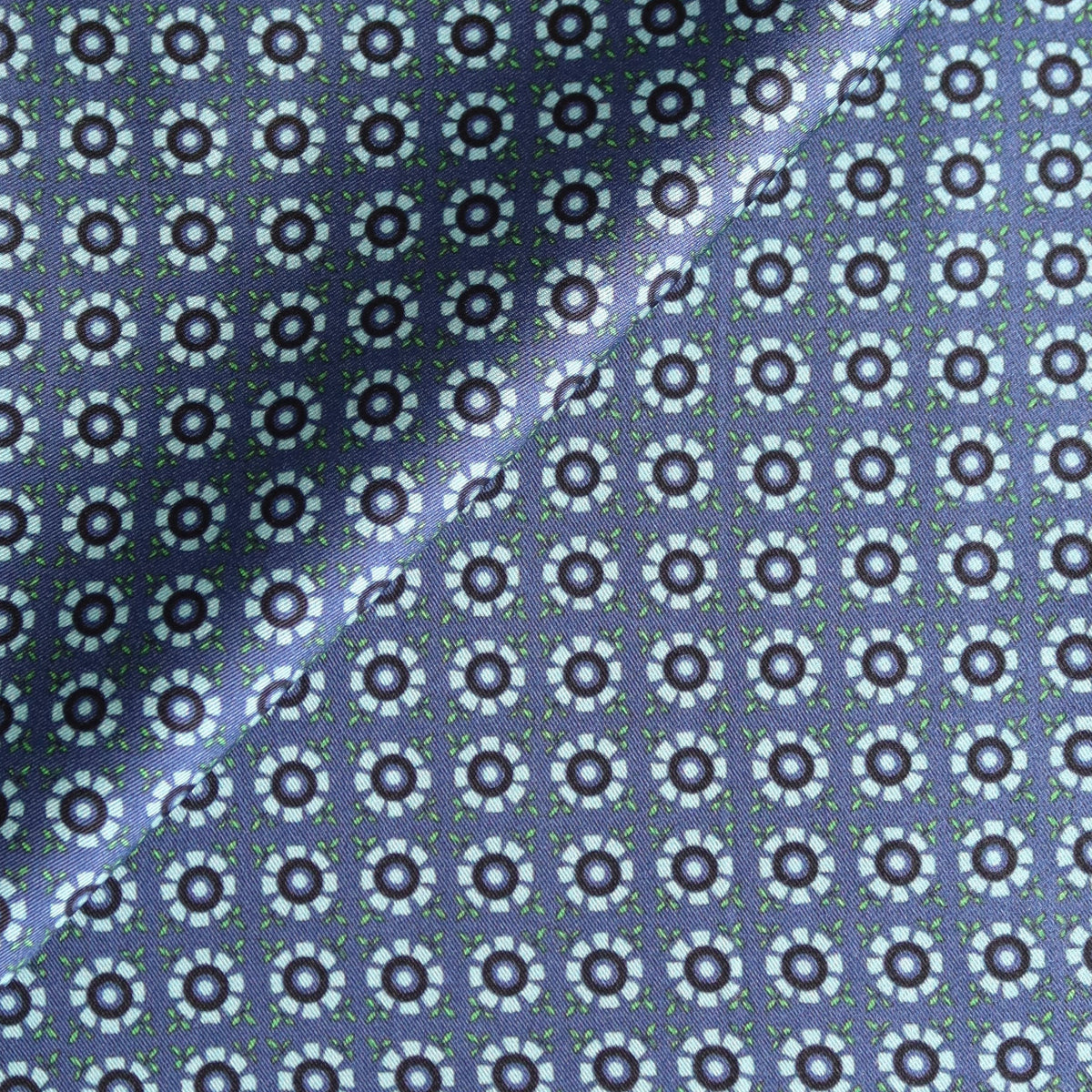 A close-up of a folded square pocket square with a dark navy border and a repeating white geometric floral pattern.