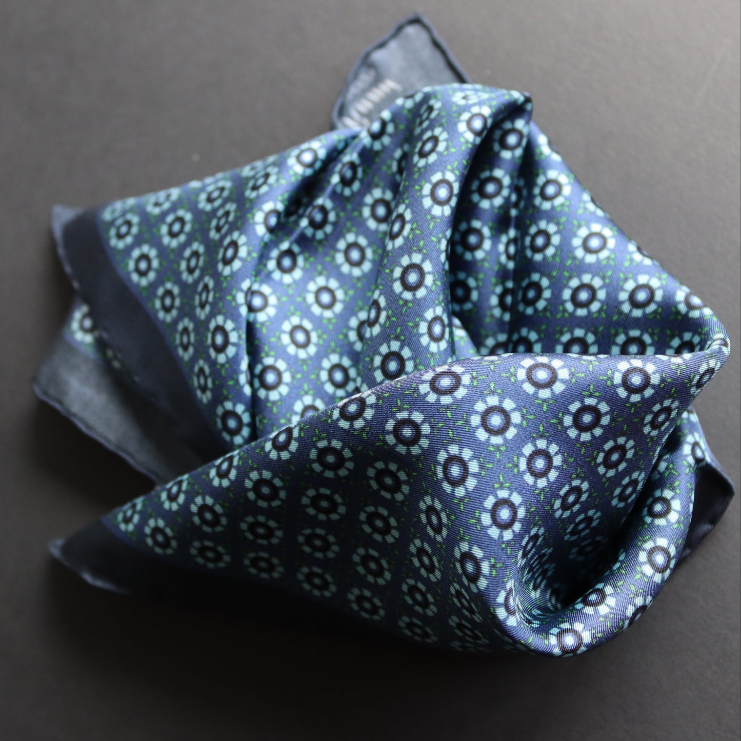 A folded blue patterned silk pocket square with a repeating white and light blue floral design, displayed on a dark grey background.