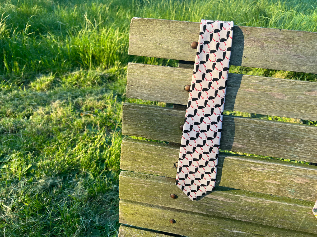 A patterned silk necktie featuring a repeating pattern of black herons with red beaks on a dusky grey base, draped over a weathered wooden bench in an outdoor grassy setting.