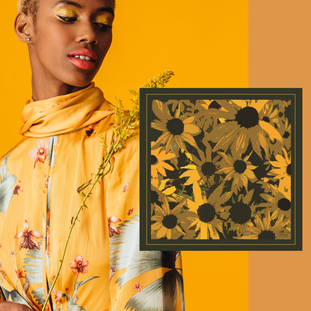 A model in a yellow floral scarf and matching top stands against a warm yellow-orange background, next to a square framed print featuring Rumi London's Gloriosa Daisy Luxury Silk Scarf.