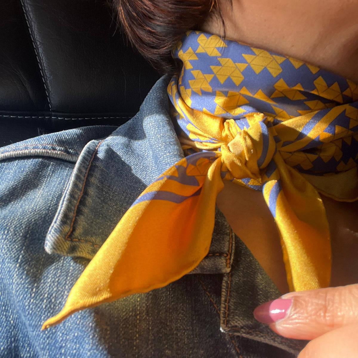 A close-up lifestyle shot of a woman wearing a yellow and blue motif patterned silk scarf tied around their neck, paired with a blue denim jacket.