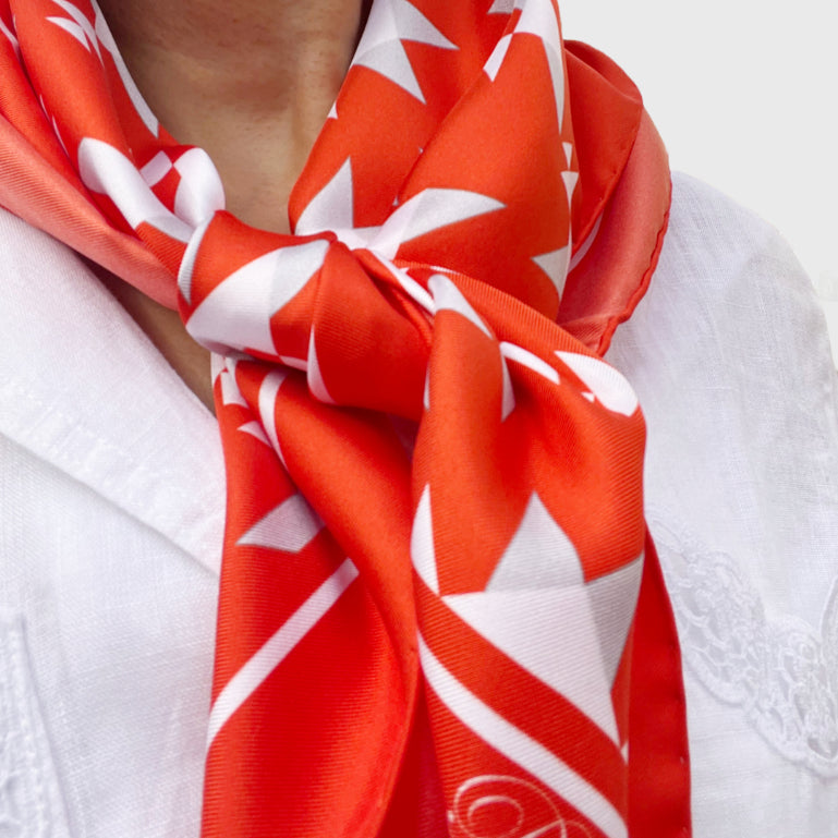 Woman wearing square silk scarf with white geometric pattern on an orange background and border, around her neck.