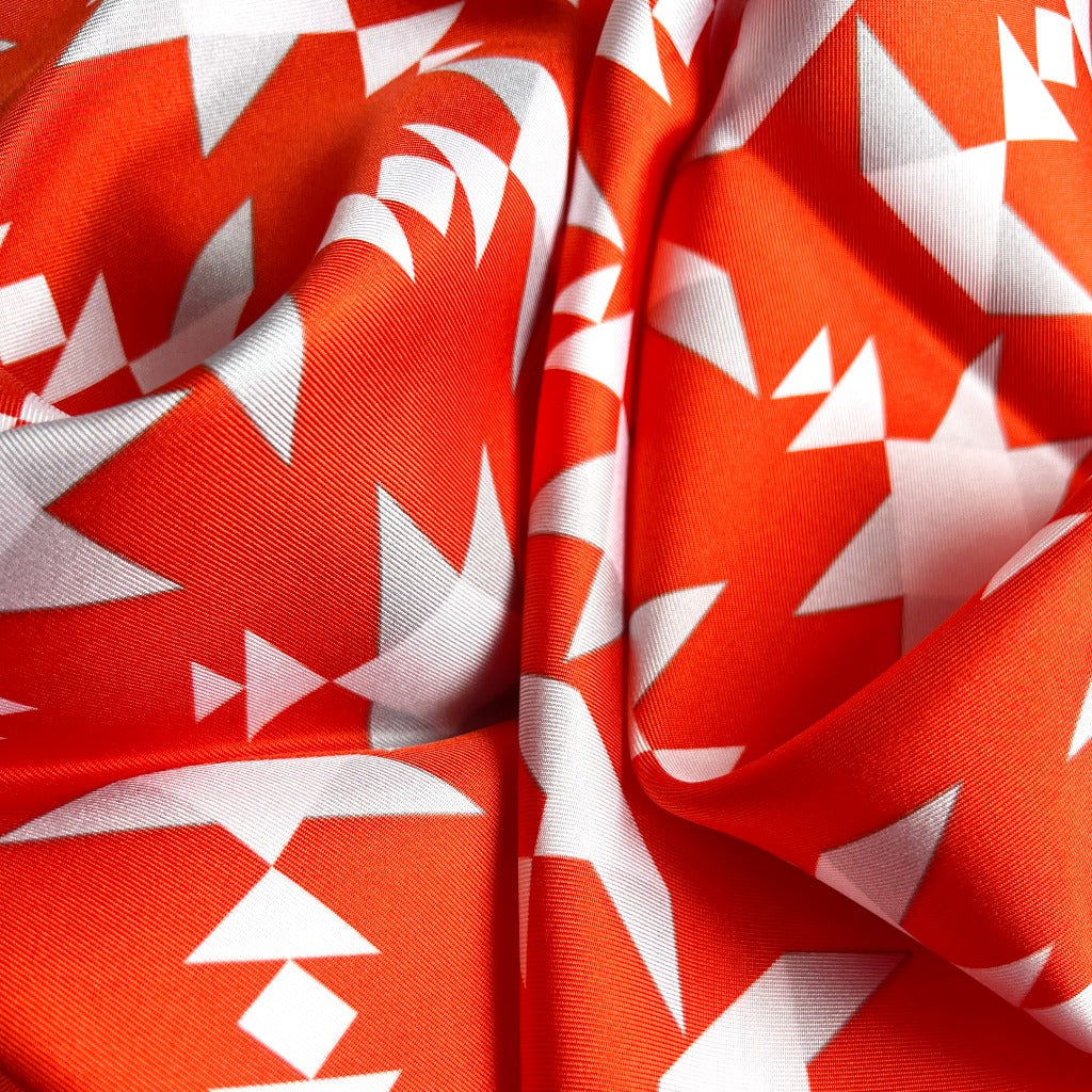 Close-up of square silk scarf with white geometric pattern on an orange background and border.